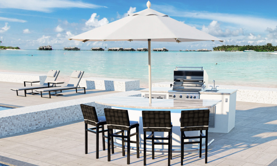 A sleek outdoor kitchen by the beach, featuring the ST. CROIX 8 + GX8 modern grill under a large white umbrella. Four black bar stools are set around a curved counter. Sun loungers rest on a white stone patio nearby, with clear turquoise waters and overwater bungalows in the background.
