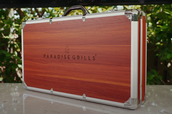 A silver and wooden case with chrome accents, labeled "Rotisserie Conversion," sits closed on a marble surface. The background features greenery. The case appears sleek and sturdy, suggesting it is intended for high-quality or specialized grilling equipment.