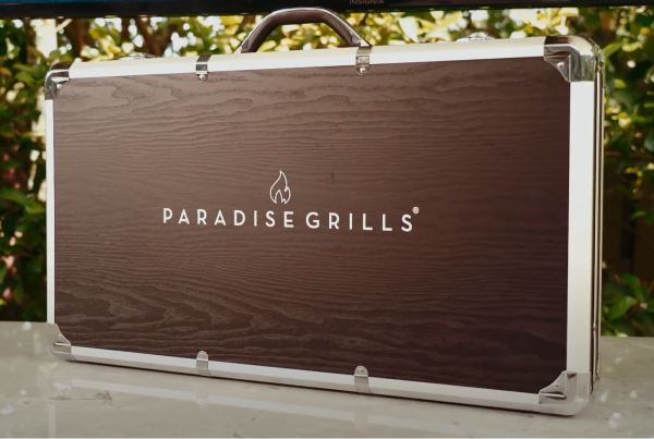The Rotisserie Conversion case is brown with silver edge accents and a handle on the top. "PARADISE GRILLS" text, along with a flame logo, is displayed in white at the center of the case. The backdrop includes blurred greenery, hinting at an outdoor setting.
