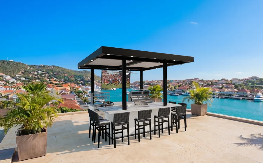 The St. Barth (Copy) features a modern rooftop kitchen and bar with black chairs, grill, TV, and palm trees, overlooking a coastal town with blue waters and hills under a clear sky.